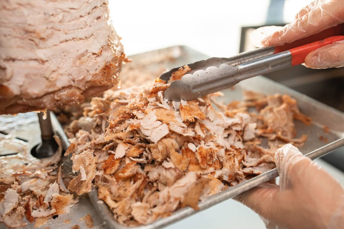Gloved hands lifting shaved doner meat with red-handled tongs onto a steel sheet tray, with the vertical spit visible at the left edge