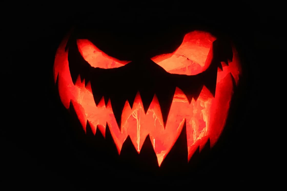 A carved jack-o'-lantern with jagged teeth and slanted eyes glows orange against total darkness on Halloween night.