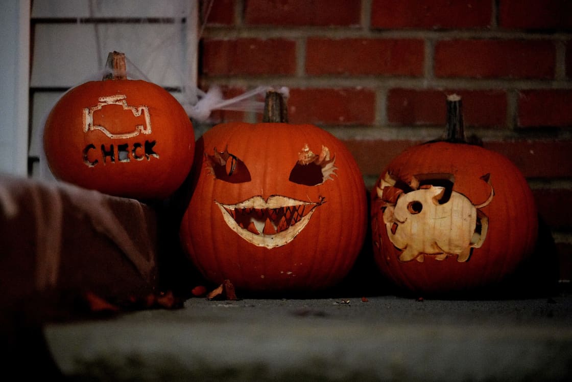 Three carved jack-o'-lanterns lit on a brick porch step at dusk, one cut with a grinning toothy face, one with a small animal shape, cobwebs strung across the window behind them