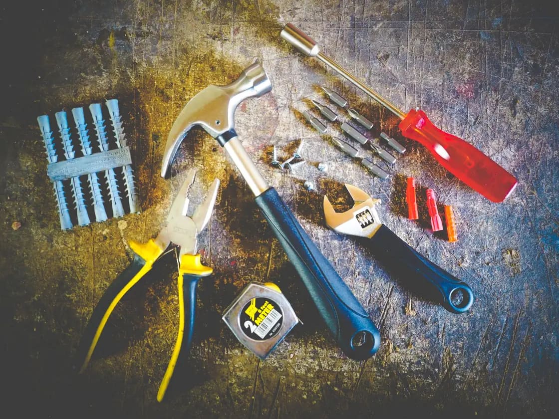 Hand tools laid out on a scratched metal surface — a hammer and adjustable wrench beside pliers, screwdriver bits and plastic wall anchors