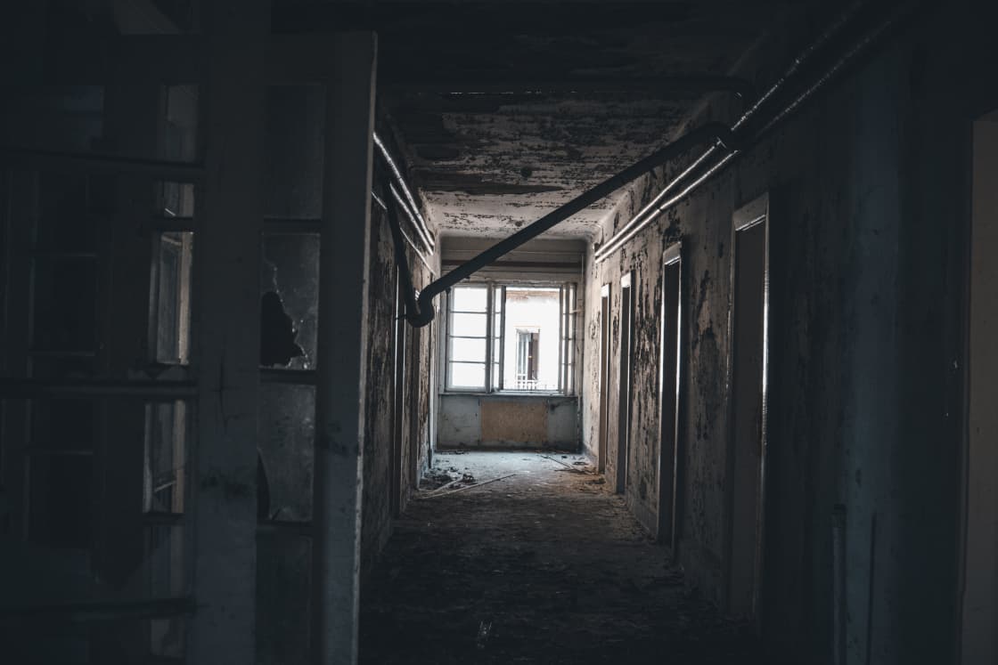 A derelict corridor with peeling plaster walls and exposed pipes running along the ceiling, dark open doorways down both sides, debris across the floor, and a bright window at the far end