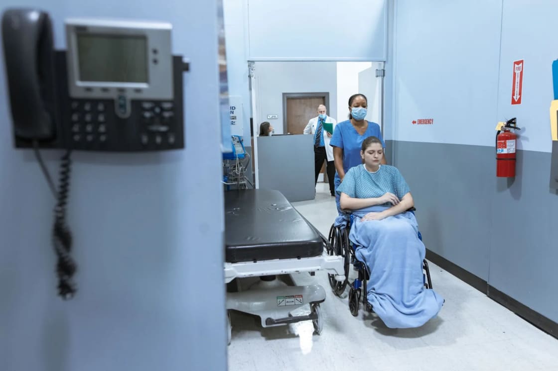 A hospital corridor with a wall-mounted telephone in the foreground and a member of staff attending a patient seated in a wheelchair further down