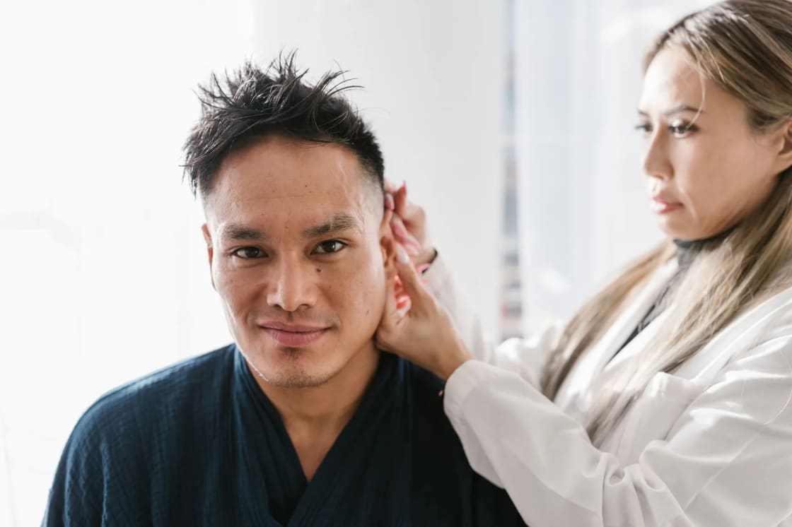 A clinician in a white coat examining the ear of a seated man in a bright consulting room