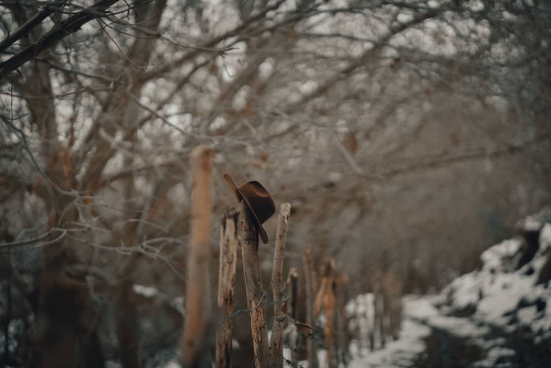 A brown felt cowboy hat hooked over a weathered wooden fence post, a line of rough posts and barbed wire running away past it into bare winter trees and patchy snow, in muted brown and gray light