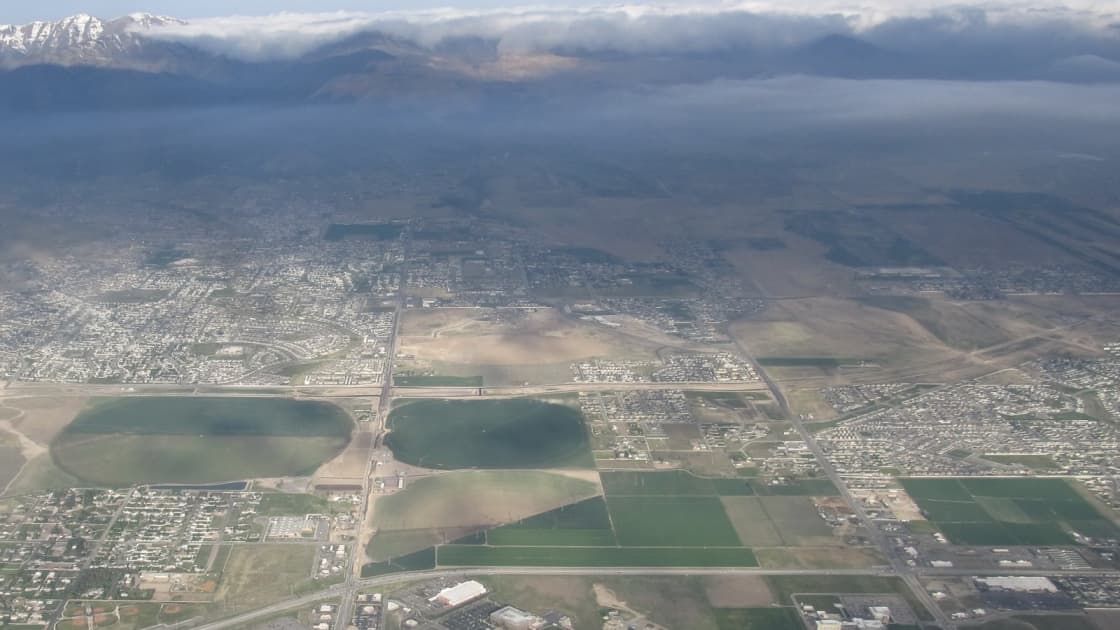 Seen from an airplane window, round irrigated fields, farmland and new neighborhoods of Riverton and Herriman spread across the valley floor, with hazy mountains and a bank of cloud on the horizon