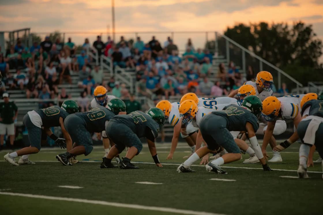 Two high-school teams set at the line of scrimmage at sunset, the home grandstand full of spectators behind them under an orange sky