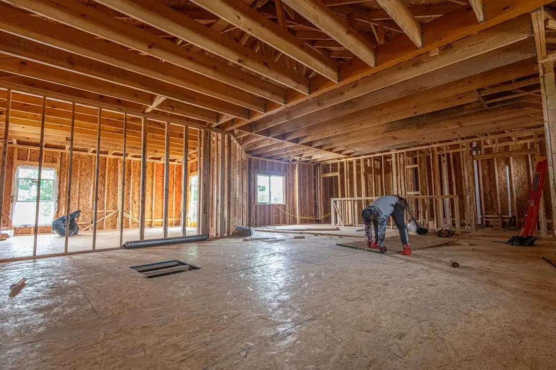 The inside of a house under construction, with unclad stud walls, exposed roof trusses overhead, an OSB subfloor and a worker crouching near installed windows