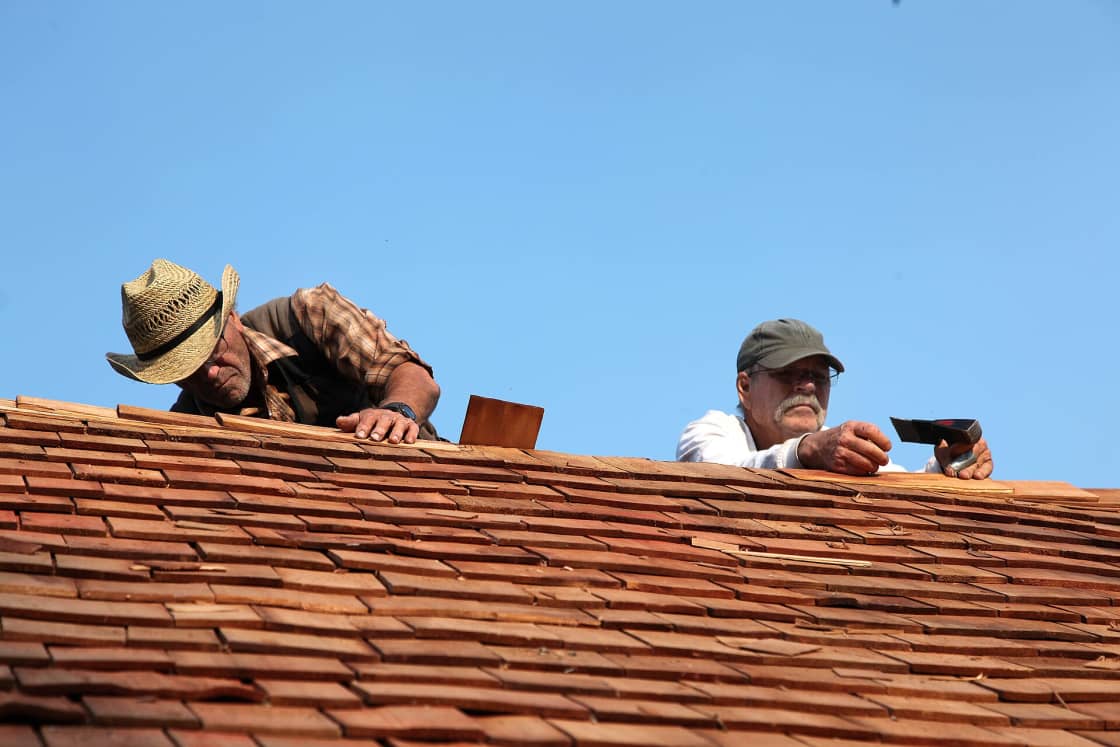 Two workers leaning over the ridge of a tile roof examining the tiles closely against a clear blue sky