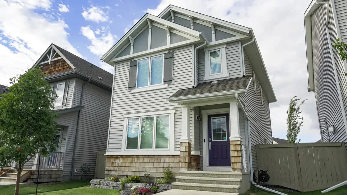 A two-story house with pale gray lap siding, white gable trim, shingle accent panels and a dark purple front door at the top of a short flight of steps, with a neighboring house close on the left