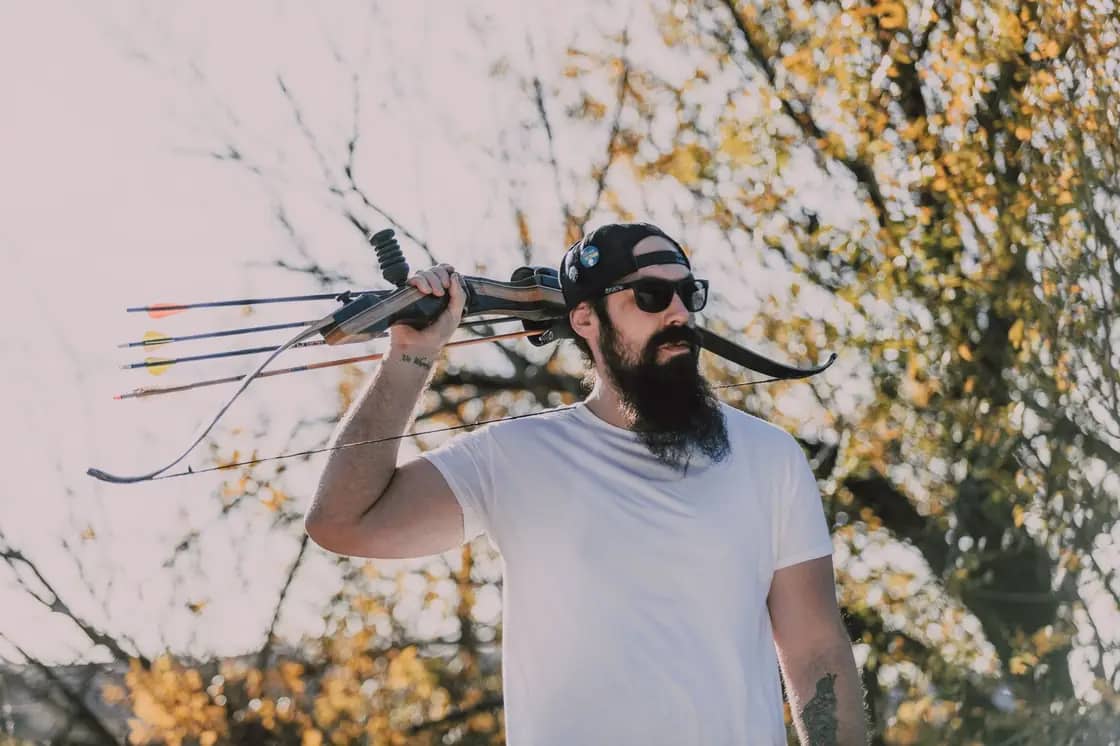 A bow hunter carrying a recurve bow and a quiver of arrows over one shoulder beneath autumn trees