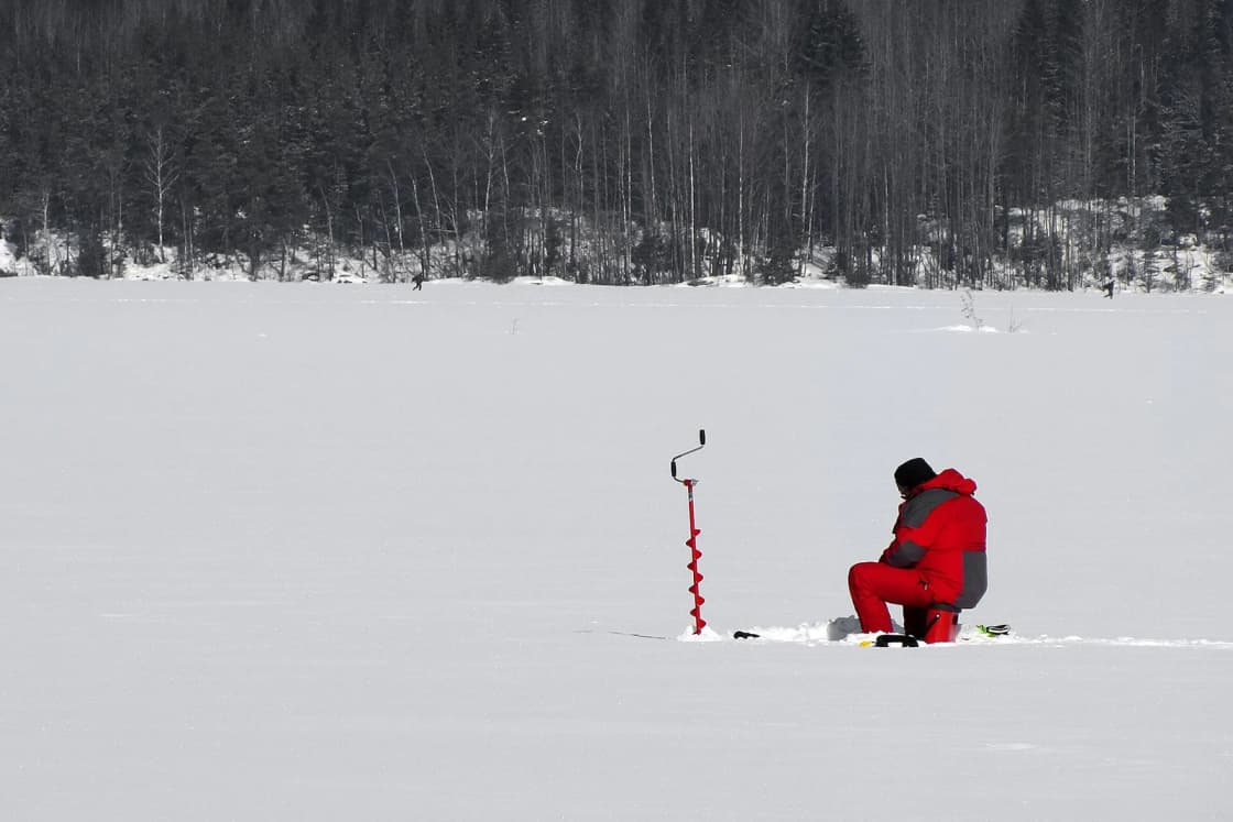 An angler in a red snowsuit sits beside an ice auger on a snow-covered frozen lake.