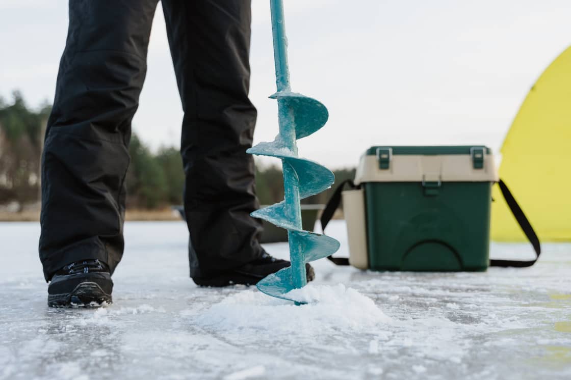 A hand ice auger standing in a drilled hole on a frozen lake beside a tackle box and the edge of a yellow shelter