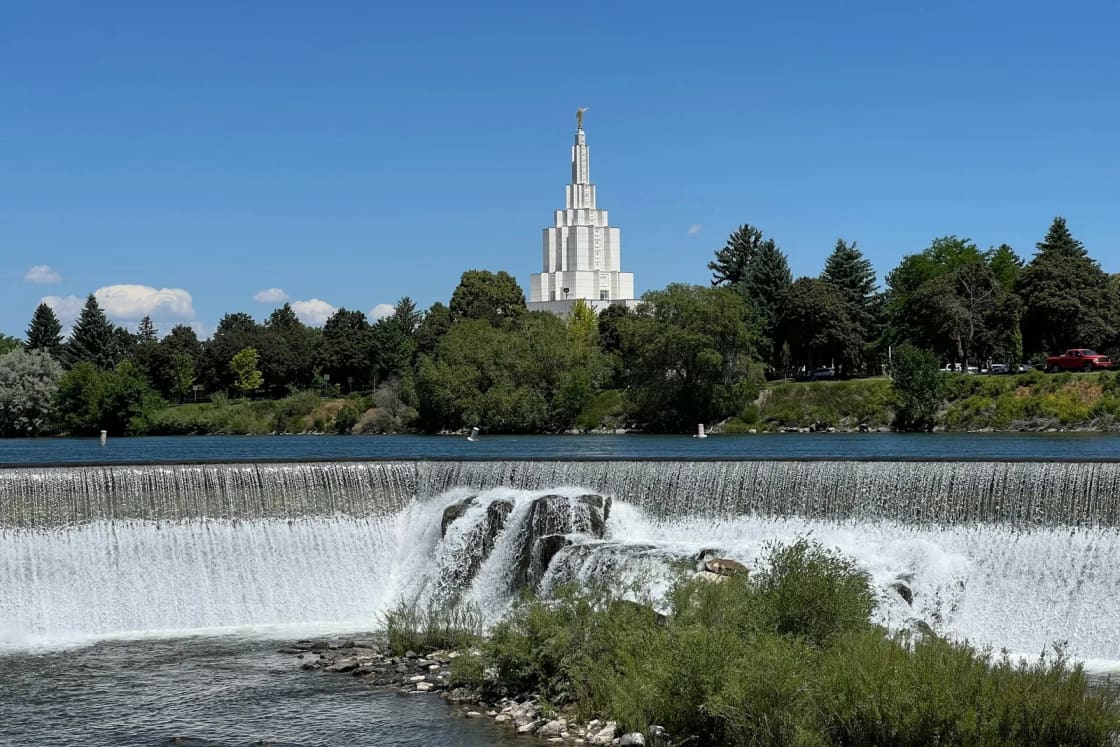 The wide falls of the Snake River at Idaho Falls spill over a long weir along the greenbelt, the white Idaho Falls temple rising above the trees.