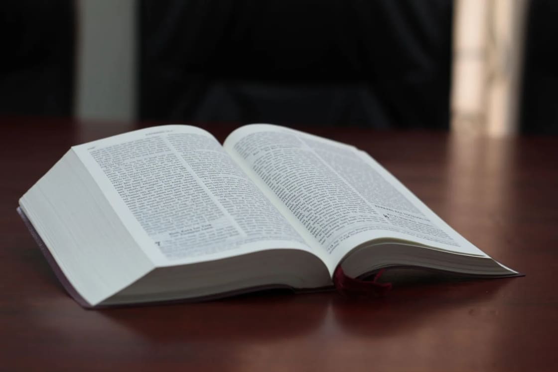 An open book with thin printed pages resting on a dark polished table, lit from one side against a black background