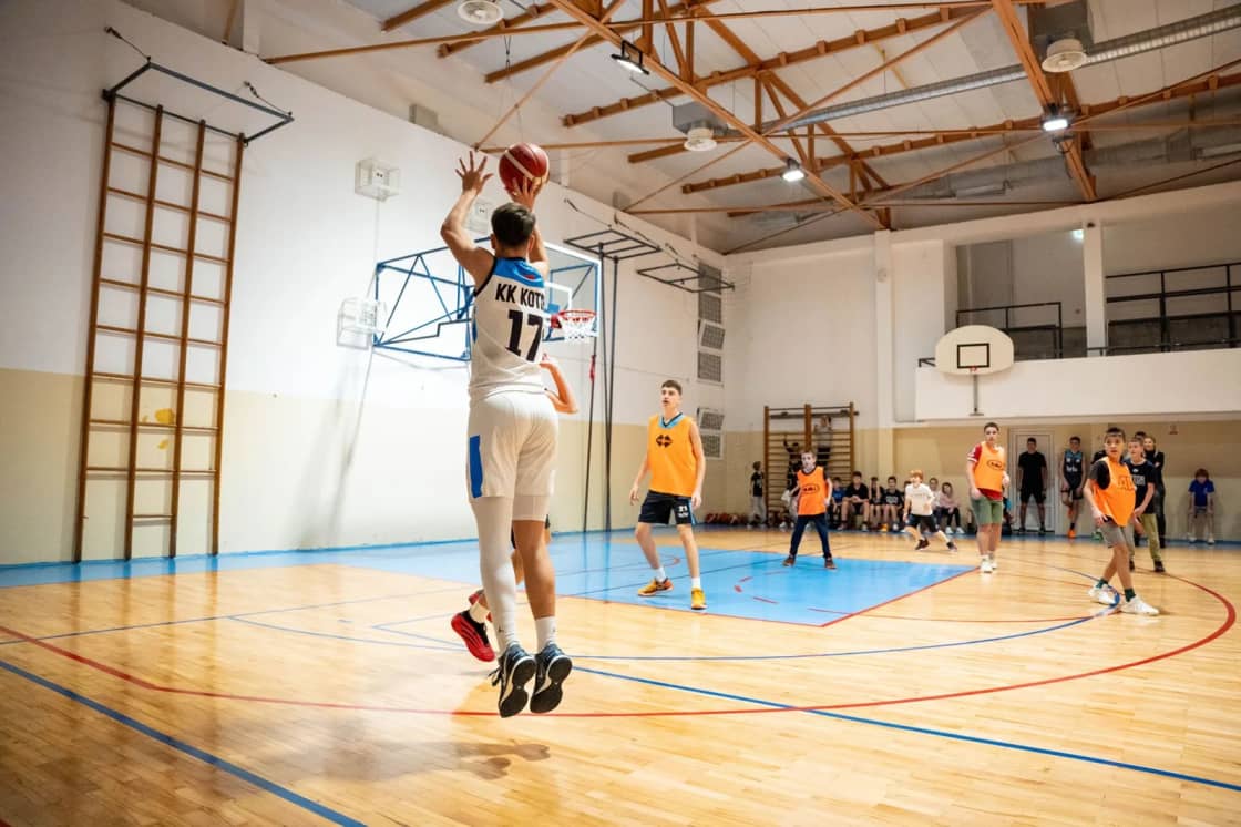 A player in a white jersey rises for a jump shot in a school gym with a blue-painted key while teammates and defenders in orange watch — youth and recreational basketball season in Utah Valley