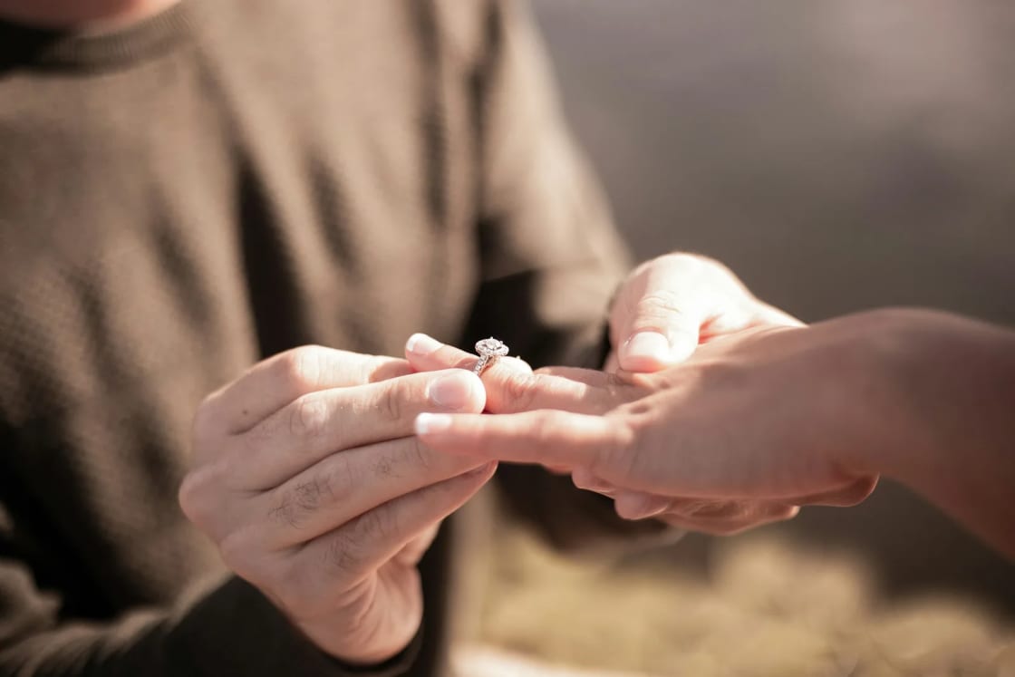 A man in a brown sweater slips a diamond engagement ring onto a woman's finger, their faces out of frame in soft outdoor light.