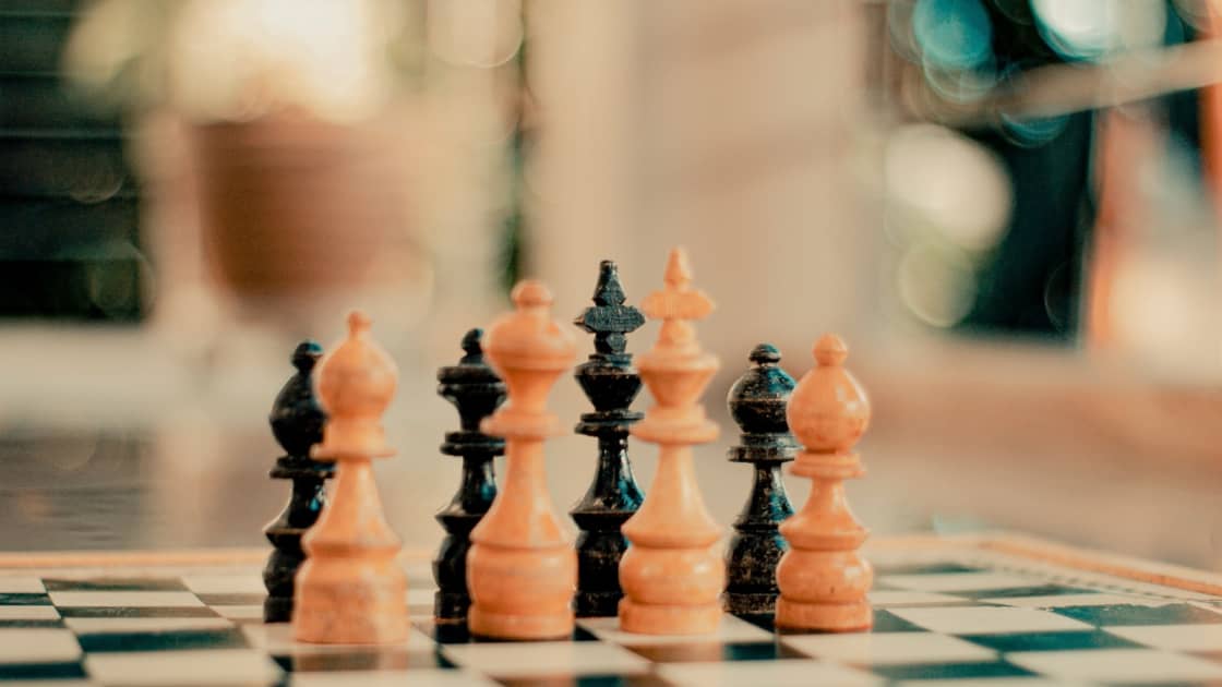 Light and dark wooden chess pieces lined up on a chessboard, with a softly blurred room behind