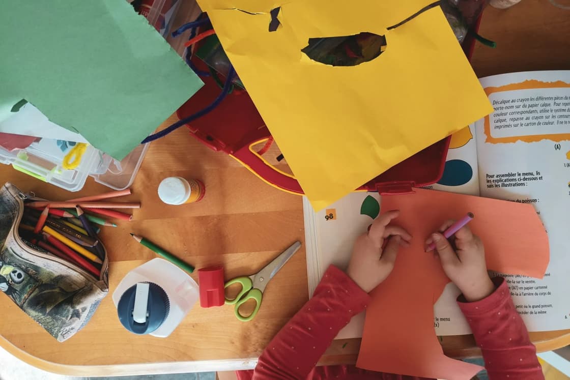 Seen from above, a child cuts and colors construction paper at a wooden table covered with scissors, glue, crayons and scraps.