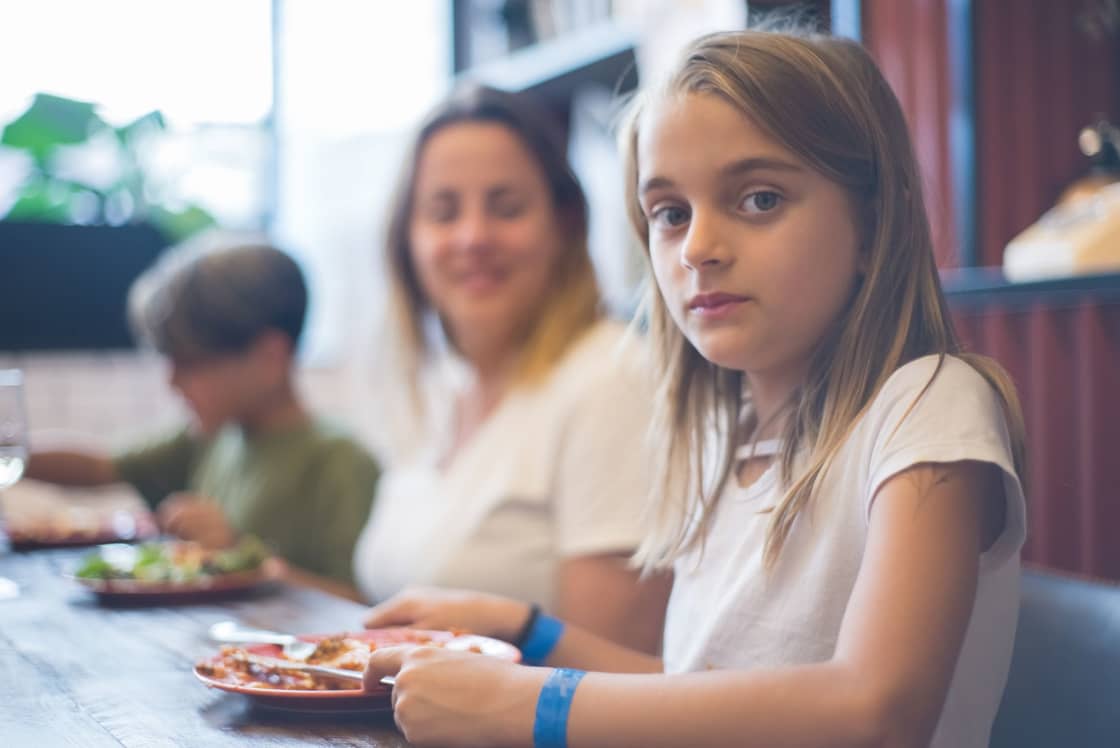 A girl at a restaurant table with a plate of food, her mother and younger brother seated beside her in a booth