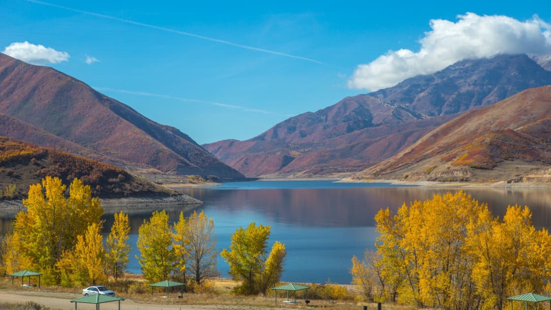 Deer Creek Reservoir under a blue sky, golden cottonwoods and green-roofed picnic shelters along the shore and russet mountains rising around the water