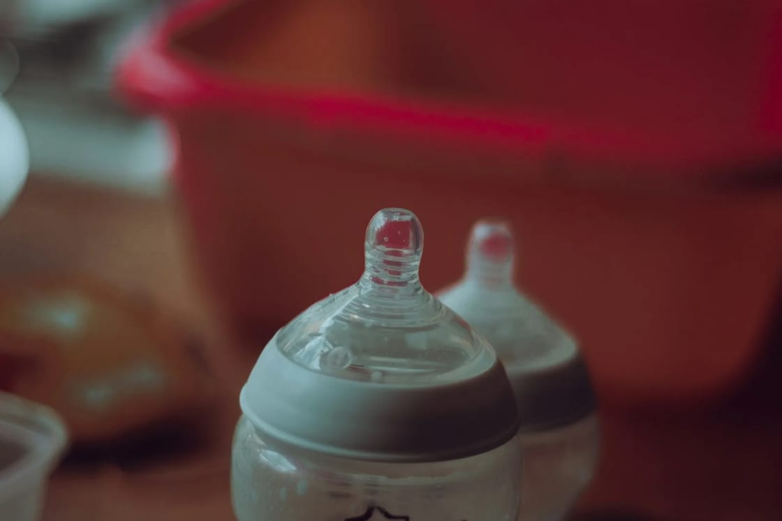 Two baby bottles with clear silicone nipples stand side by side on a table against a soft red background, ready for the next feeding.