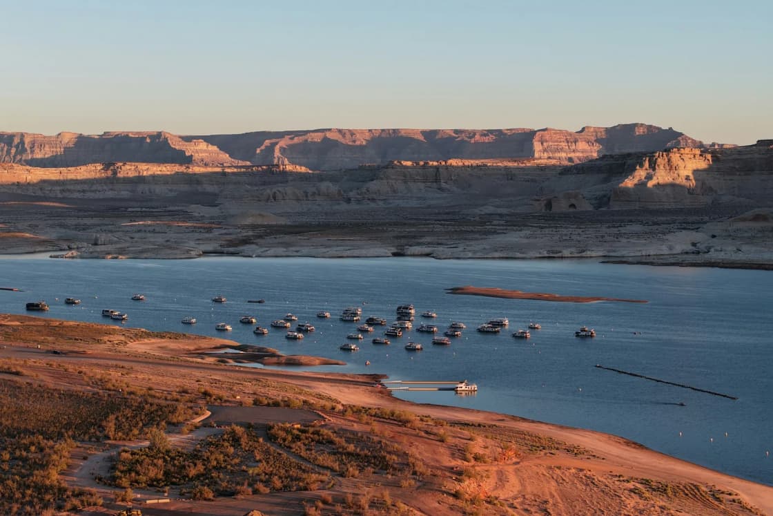 Houseboats and powerboats moored in a Lake Powell bay at sunrise, with red sandstone shoreline in front and long desert mesas behind.