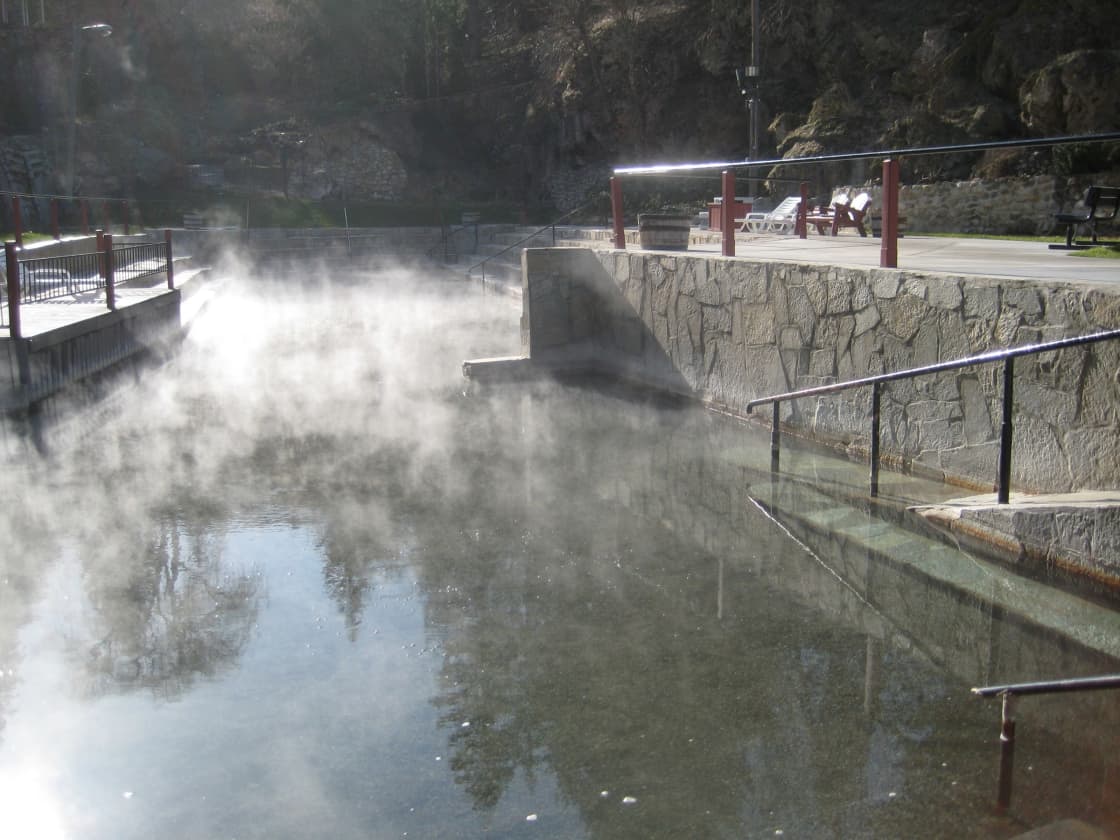Steam rising off a clear hot mineral pool at Lava Hot Springs, Idaho, with stone-faced walls, metal handrails leading down into the water and lounge chairs on the deck above