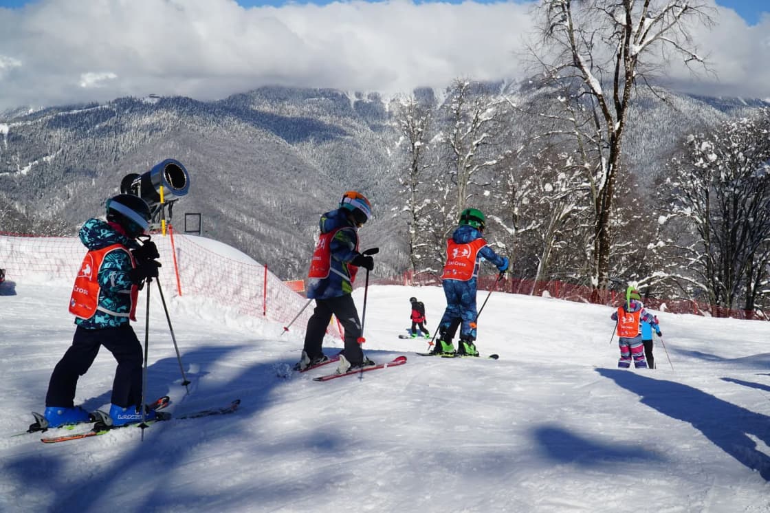 Children in helmets and bright vests stand on skis in a line beside an instructor on a sunny slope, with snowy mountains behind them