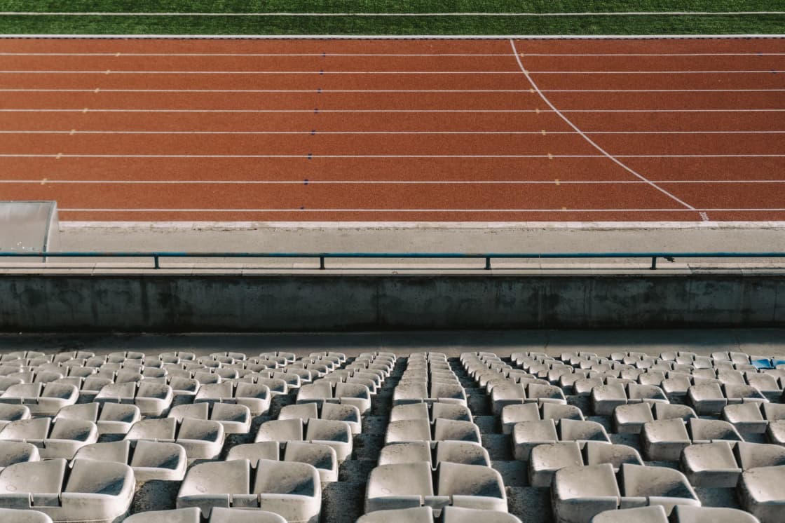 Rows of weathered pale plastic stadium seats in the foreground with a red synthetic running track and white lane lines beyond a low concrete wall