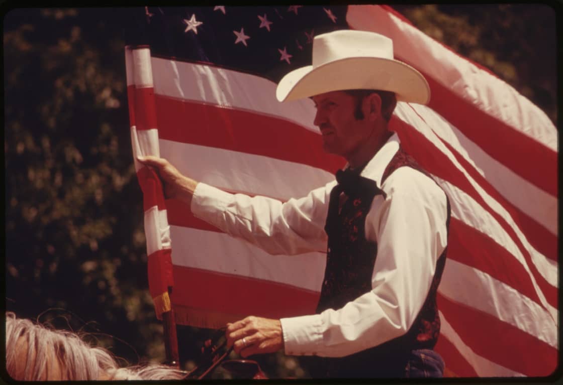 A rider in a white cowboy hat carrying an American flag on horseback during a western parade