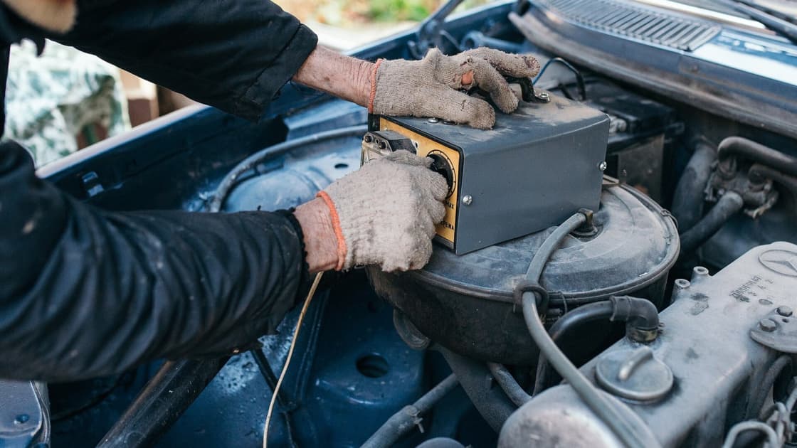 Gloved hands holding a portable diagnostic meter over the open engine bay of an older vehicle