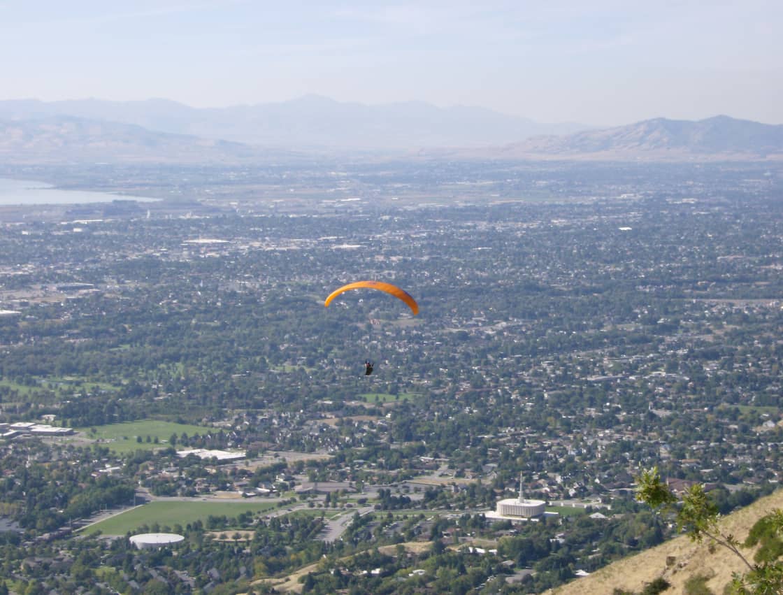 Provo, Utah seen from the mountainside above the city — a dense grid of tree-lined residential streets running west toward Utah Lake, with a paraglider over the neighborhoods