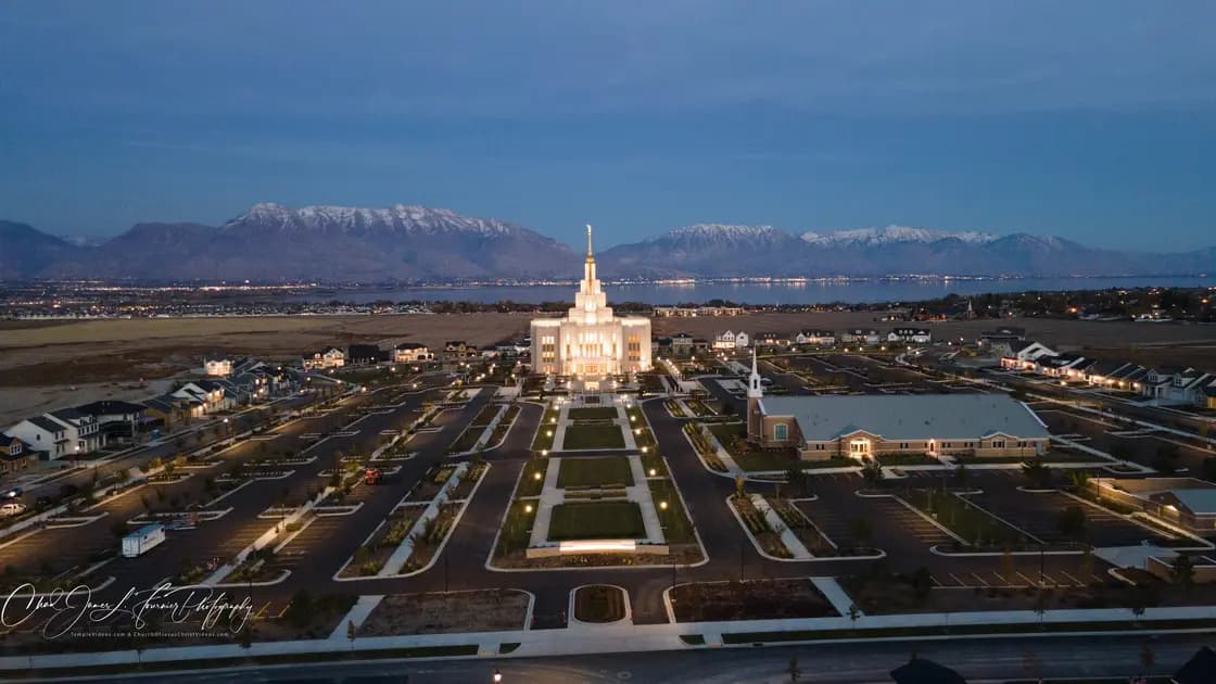An aerial view of the Saratoga Springs Utah Temple, a light stone building with a tall central spire, on its landscaped grounds