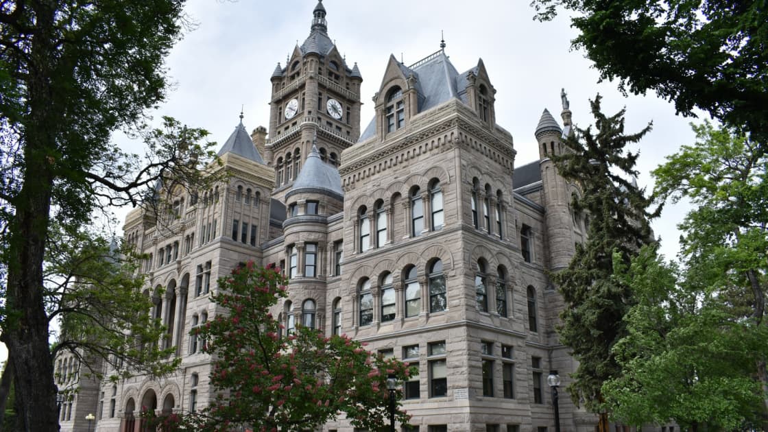 The sandstone Salt Lake City and County Building, with its clock tower, turrets and arched windows, rising behind shade trees and a flowering tree on Washington Square