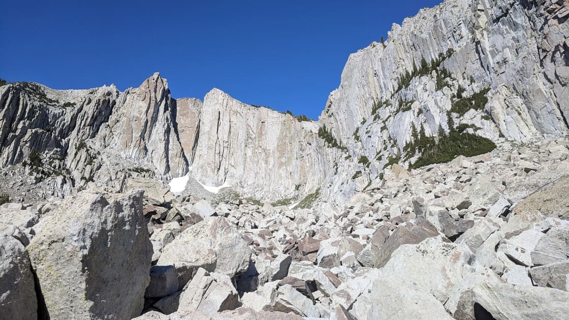 The pale granite walls of the Lone Peak Cirque rising above a field of broken boulders, with a small patch of snow at the foot of the cliffs and a deep blue sky overhead