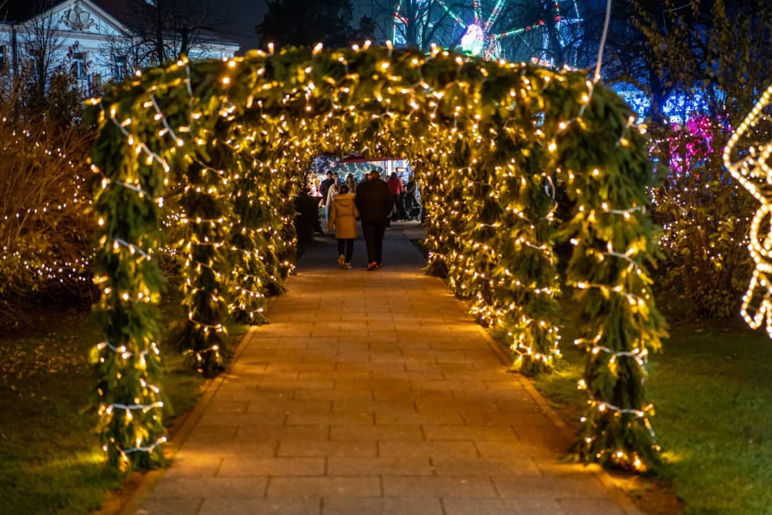 Visitors walk beneath a long tunnel of garland arches strung with warm white lights at an outdoor holiday light display after dark — the Luminaria light walk at Thanksgiving Point