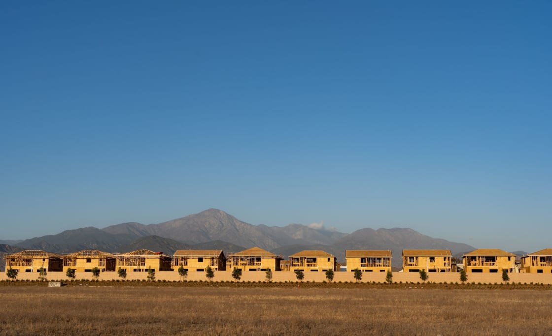 A long row of timber-framed houses under construction behind a boundary wall, with dry grassland in front and a mountain range on the horizon