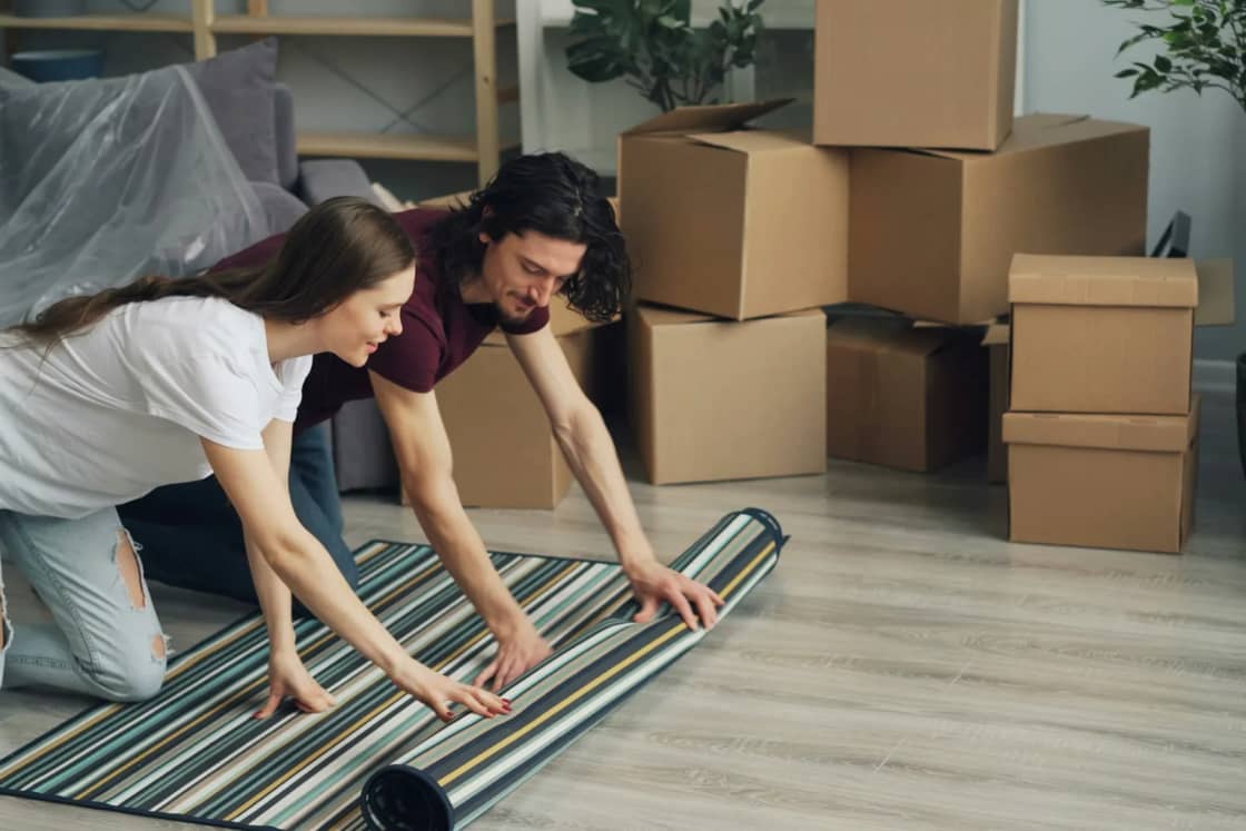 A young couple laughing as they unroll a striped rug in a new apartment, moving boxes stacked behind them