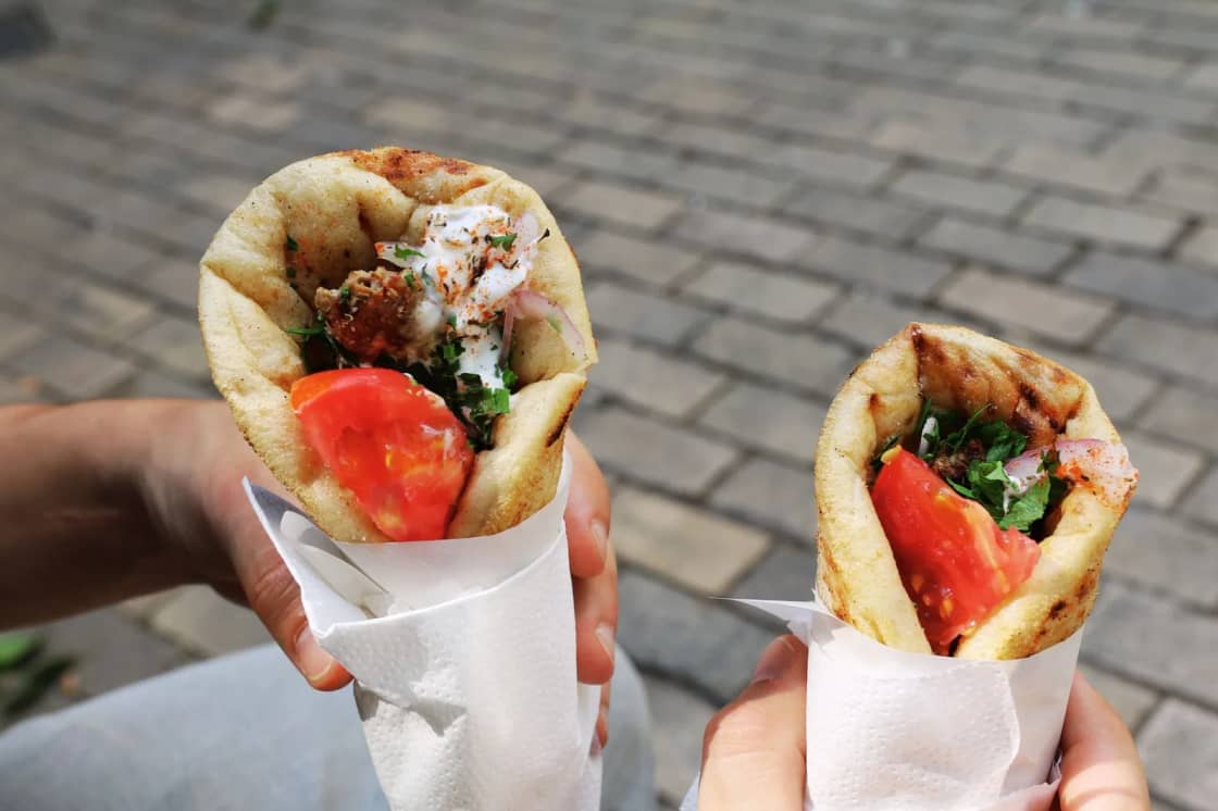 Two hands each holding a filled gyro wrapped in paper, with tomato, lettuce and white sauce visible at the open end