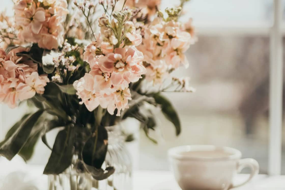 A quiet windowsill with pale pink flowers in a glass vase, a cup of tea and a small closed notebook in soft daylight