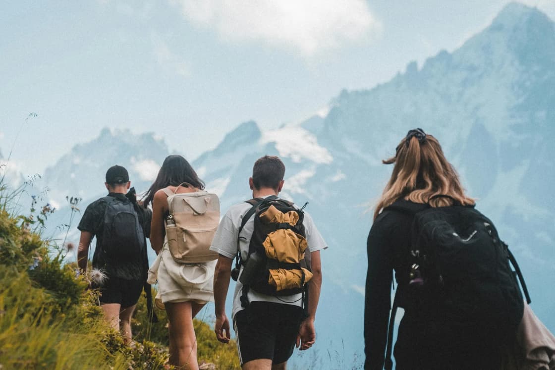 Four friends with daypacks hike single file along a grassy ridge trail toward jagged, snow-streaked peaks under puffy clouds.