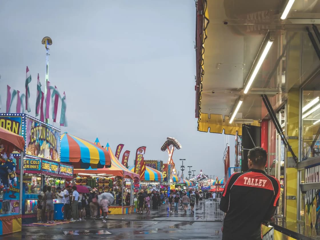 A bustling carnival with colorful tents, lively crowds, and a rainy backdrop. A man observes the festivities