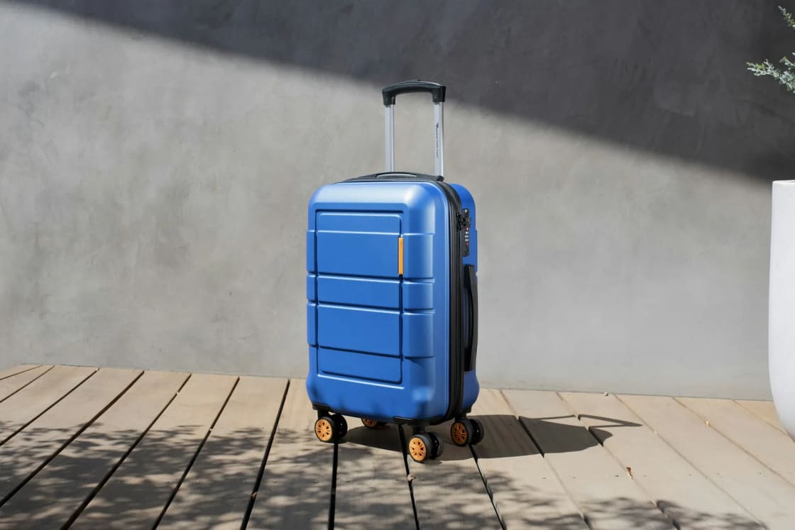 A packed blue carry-on suitcase standing on a sunny wooden deck, handle up and ready to roll