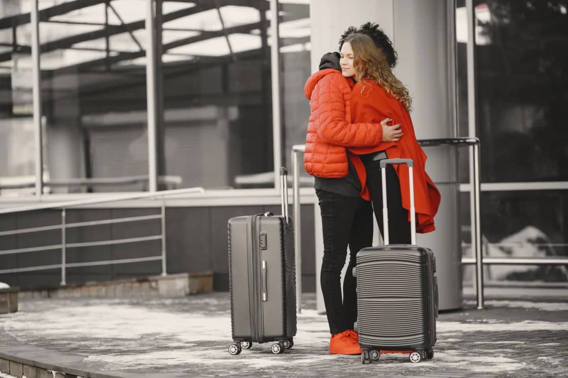 Two people embracing beside two upright wheeled suitcases on a snowy pavement outside a glass-fronted building, the red coats picked out against a desaturated background