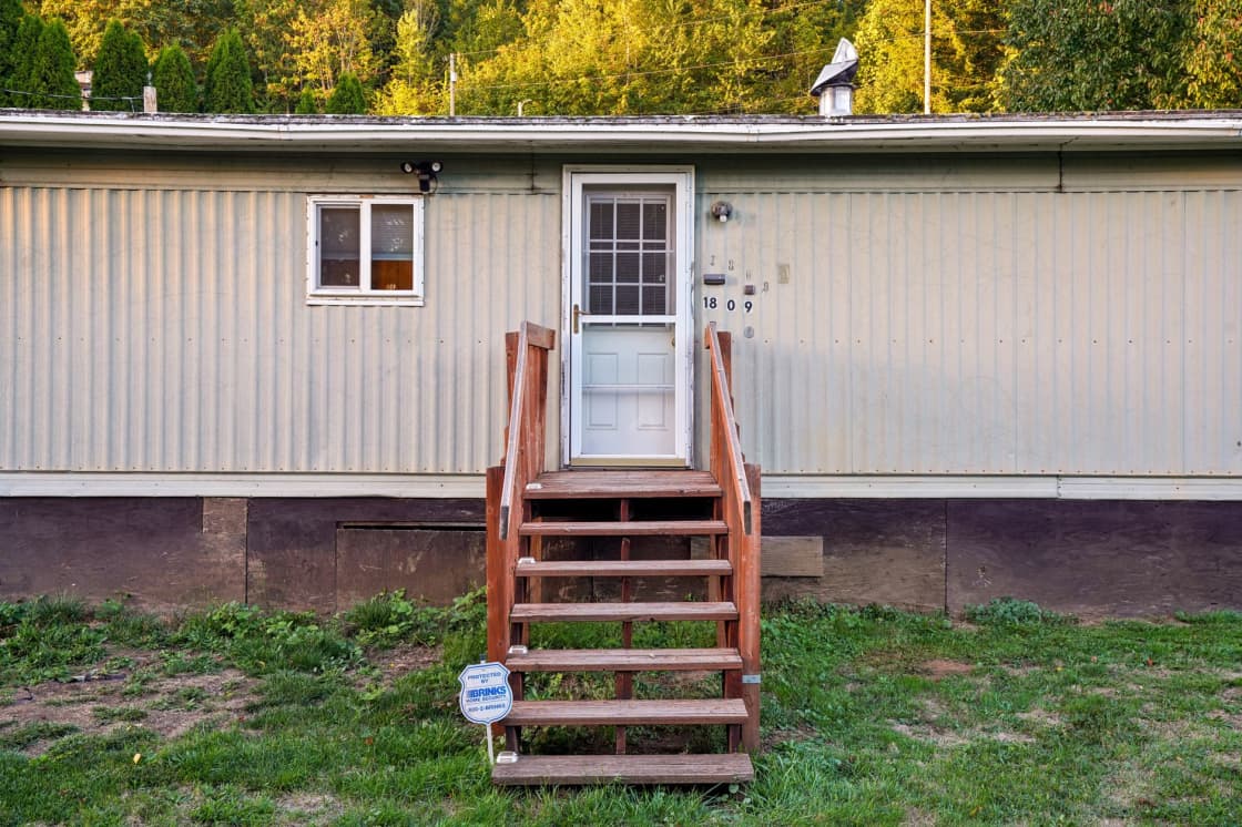 A single-wide manufactured home with pale horizontal siding, dark skirting along its base and a wooden stairway up to the front door, on a grassy lot