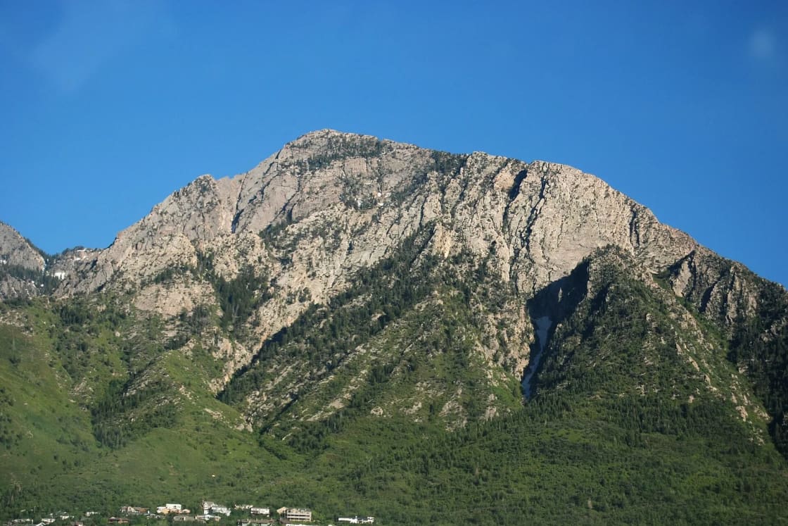 The craggy rock face of Mount Olympus rising above green foothills and the edge of the Salt Lake Valley under a clear blue sky