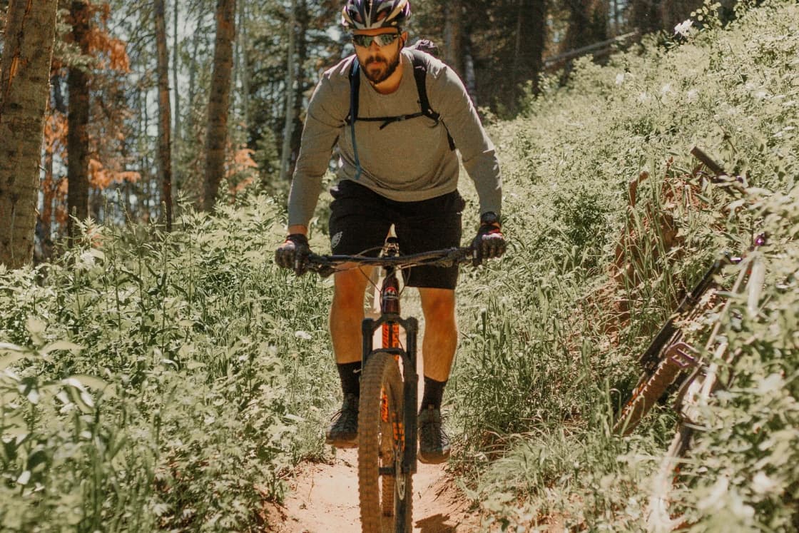A mountain biker in a helmet rides a narrow dirt singletrack between tall pines and wildflowers on a Wasatch trail