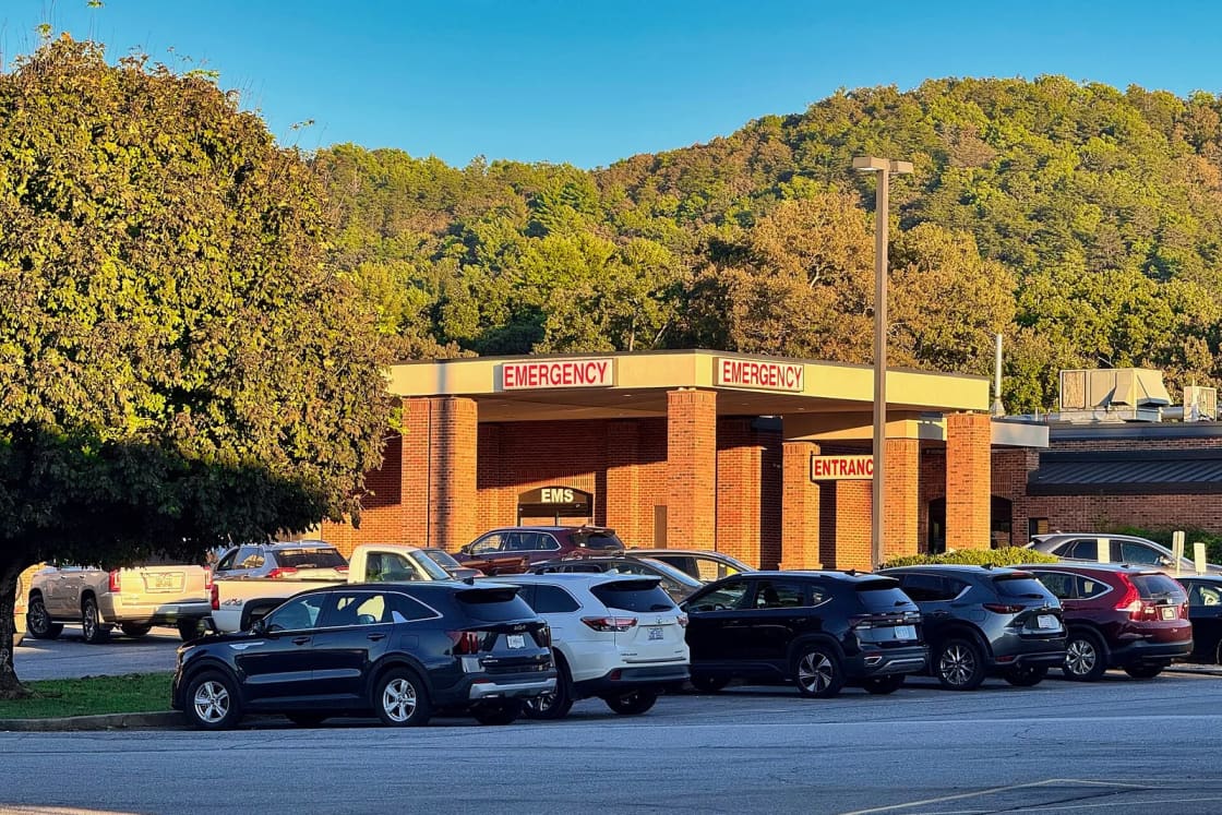 Parked cars in front of a brick hospital emergency entrance with red Emergency signs and a wooded hill behind.