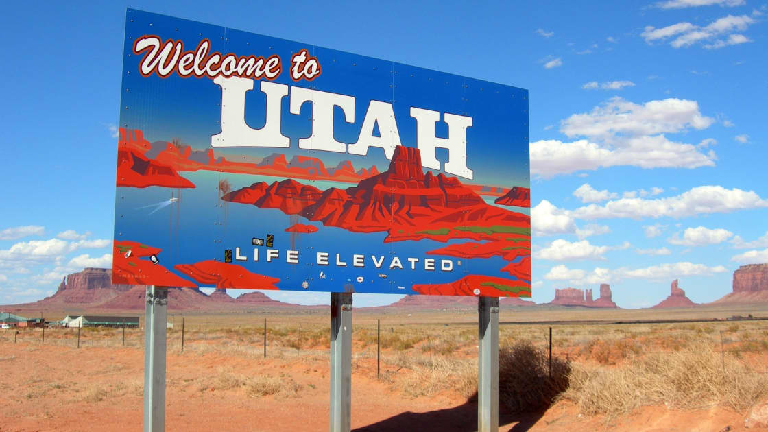 The Welcome to Utah sign beside the highway on red desert ground, with the buttes of Monument Valley on the horizon under a blue sky