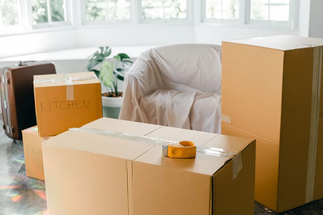 Cardboard moving boxes stand in a bright living room beside furniture draped in dust sheets and a tall window